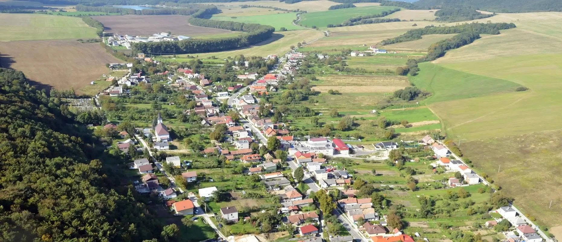 Letecký pohľad na obec Buková — domy pozdĺž hlavnej cesty, kostol, polia a lesy Malých Karpát